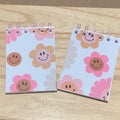 Two small notebooks with smiley face flower designs on a wooden surface