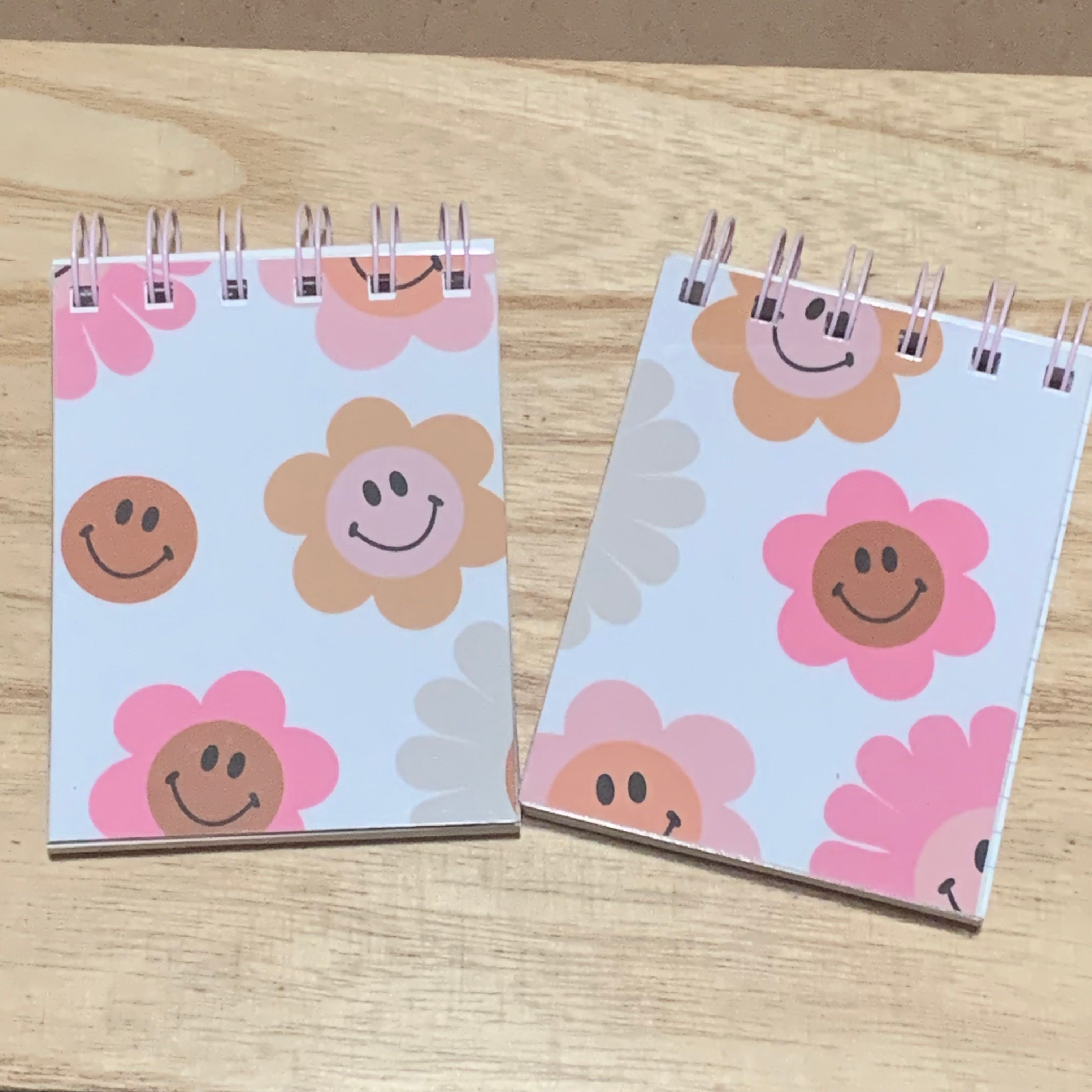 Two small notebooks with smiley face flower designs on a wooden surface
