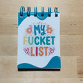 Notebook titled 'My Fucket List' on a wooden surface